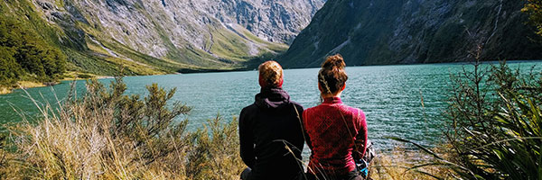 Two ladies sitting with their backs to the viewer looking at a lake and moutains.
