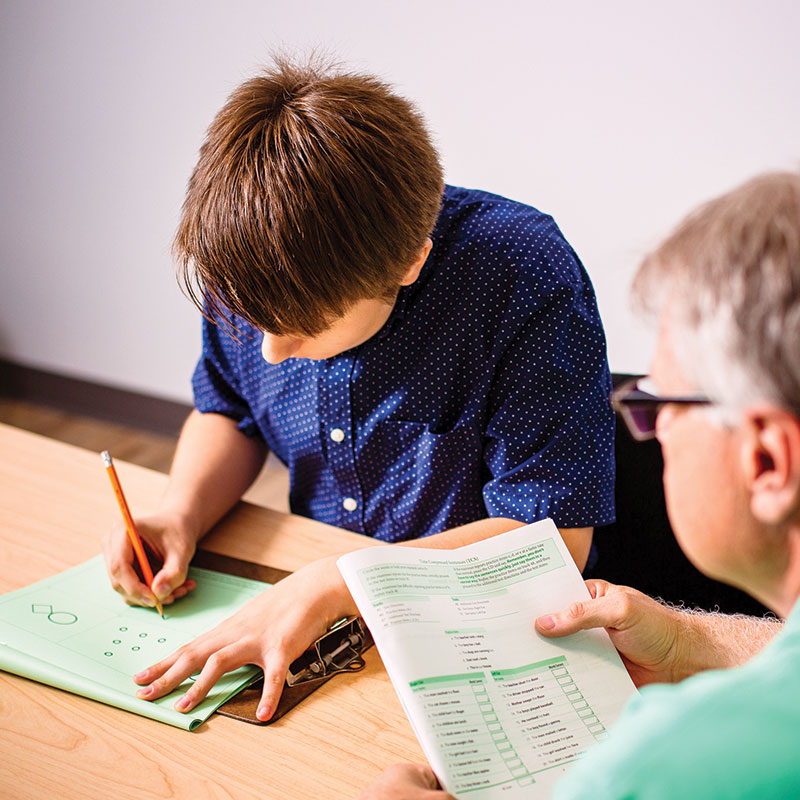 Talk Therapy A client sits at a table filling taking a writing test with a clinician on looking during a neuropsychological test.