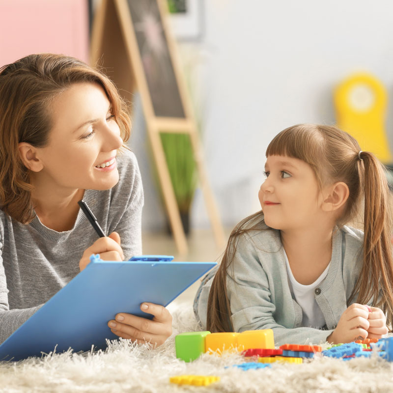 Assessments A therapist with a clipboard talks with a child on the floor demonstrating psychological testing.